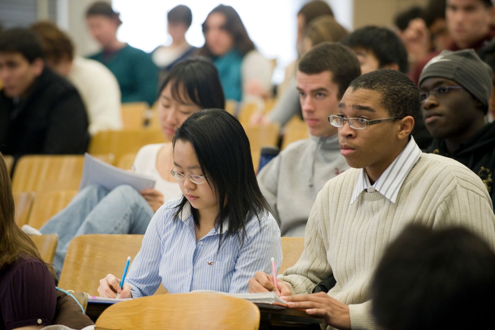 Students sit in an auditorium-style classroom. Some are taking notes while others look straight ahead.