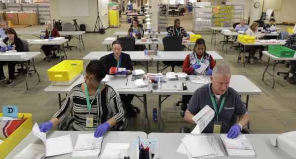 People sit two to a table, in rows facing the camera, as they process ballots.