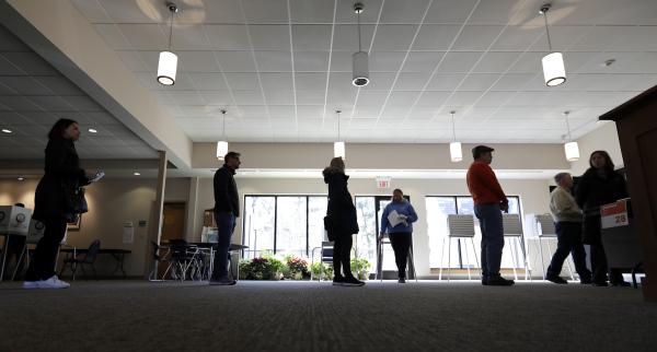 People stand in a line, backlit by windows. They are standing six feet apart from each other.