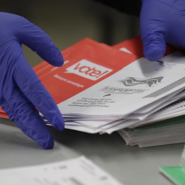 We see a pair oif hands, with purple gloves on, stacking red and white vote by mail ballots.