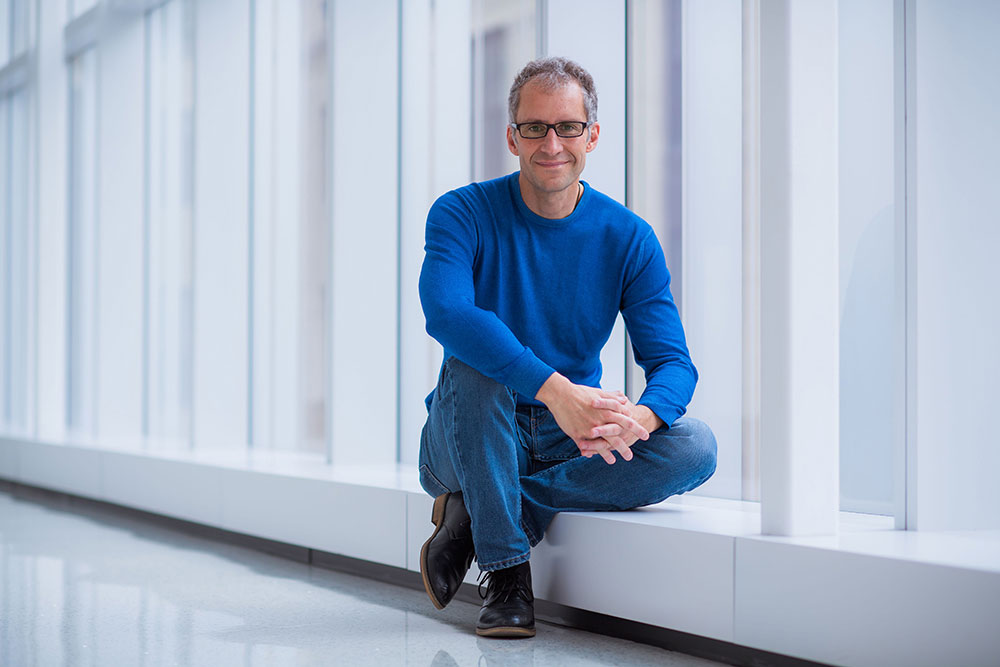 male faculty member seated in a hallway with windows along the right side, MIT