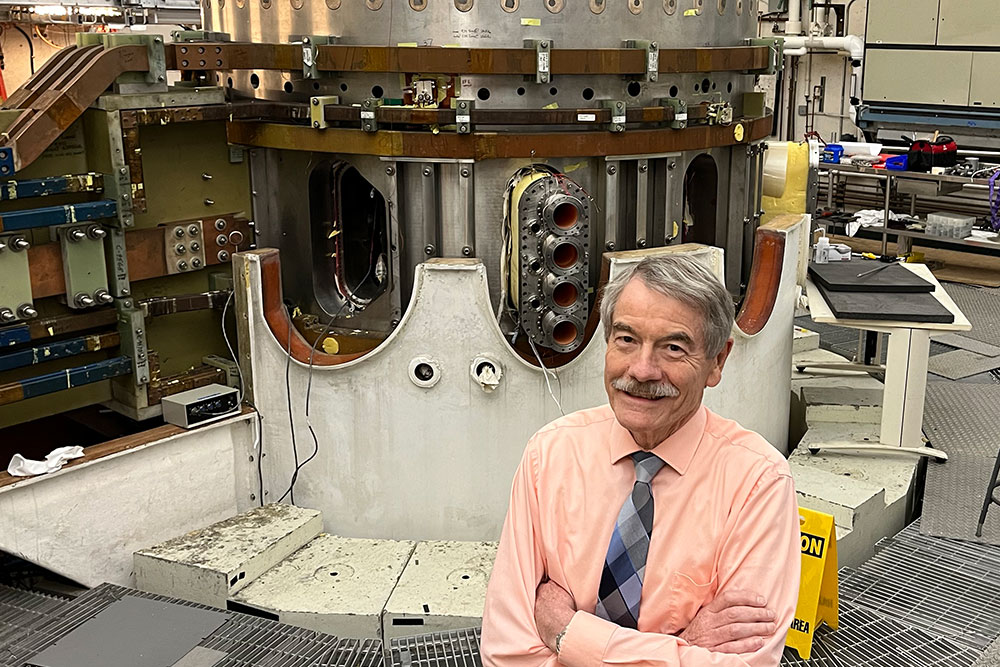 Male faculty member Ian Hutchinson in front of Alcator C-Mod, a fusion tokamak in a large lab sapce, MIT