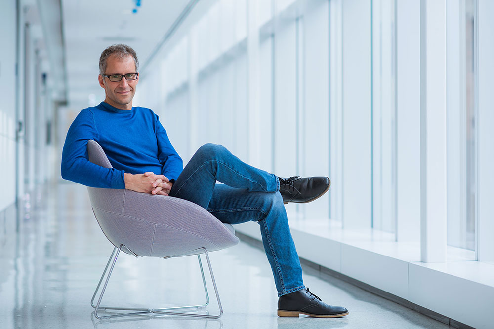 Male faculty member in blue sweater seated in a chair in a hallway, MIT