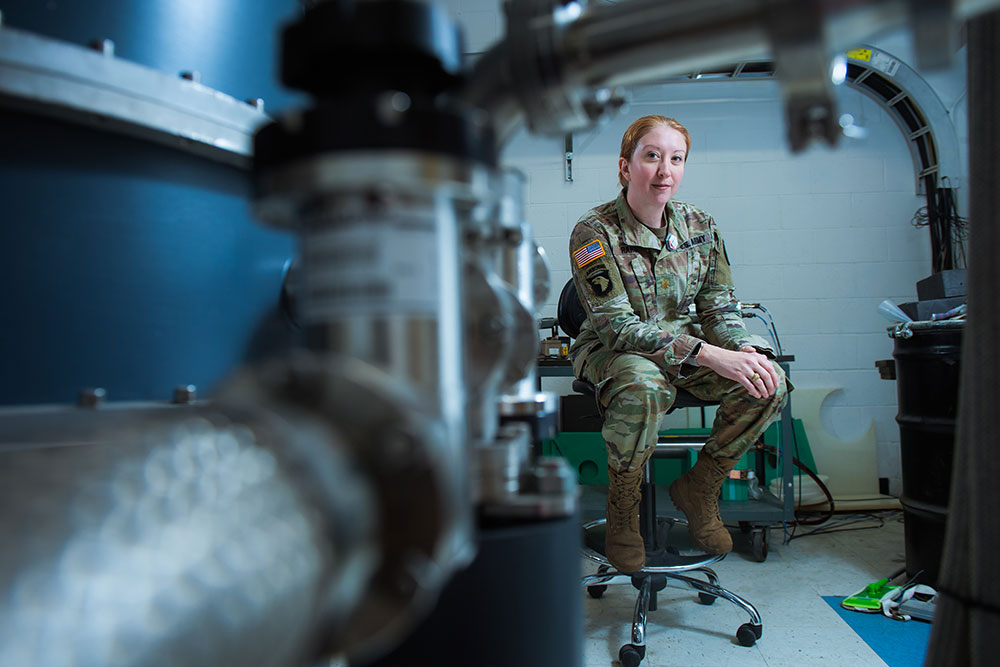 Jill Rahon in Army fatigues seated on a swivel chair in a lab with instuments and lab equipment behind her, and our of focus to the front left, MIT