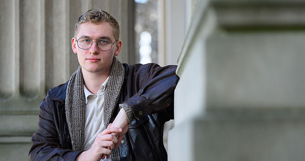 Graduate student, Liam Jines, seated outdoors with concrete architectural columns in the background and to the right